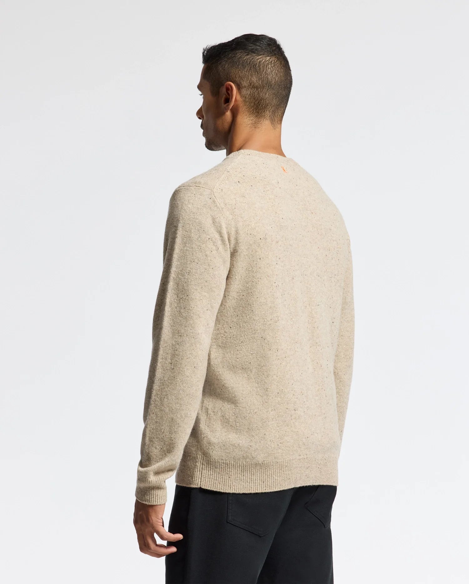 A person with short dark hair is standing and facing away, wearing a MENS LINCOLN MERINO WOOL SWEATER by Psycho Bunny in light beige, paired with dark pants against a plain white background.