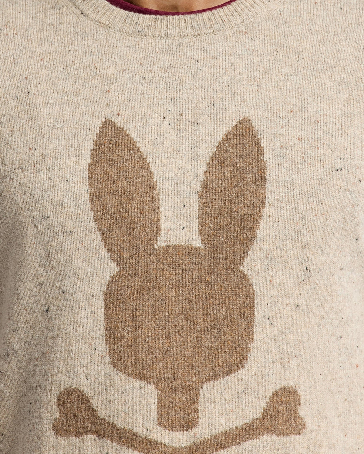 The MENS LINCOLN MERINO WOOL SWEATER - B6E831E200 by Psycho Bunny is showcased in a close-up image. This beige sweater features a brown silhouette of a stylized rabbit head with long ears above two crossed bones, highlighting the texture of the warm Merino wool knit fabric.