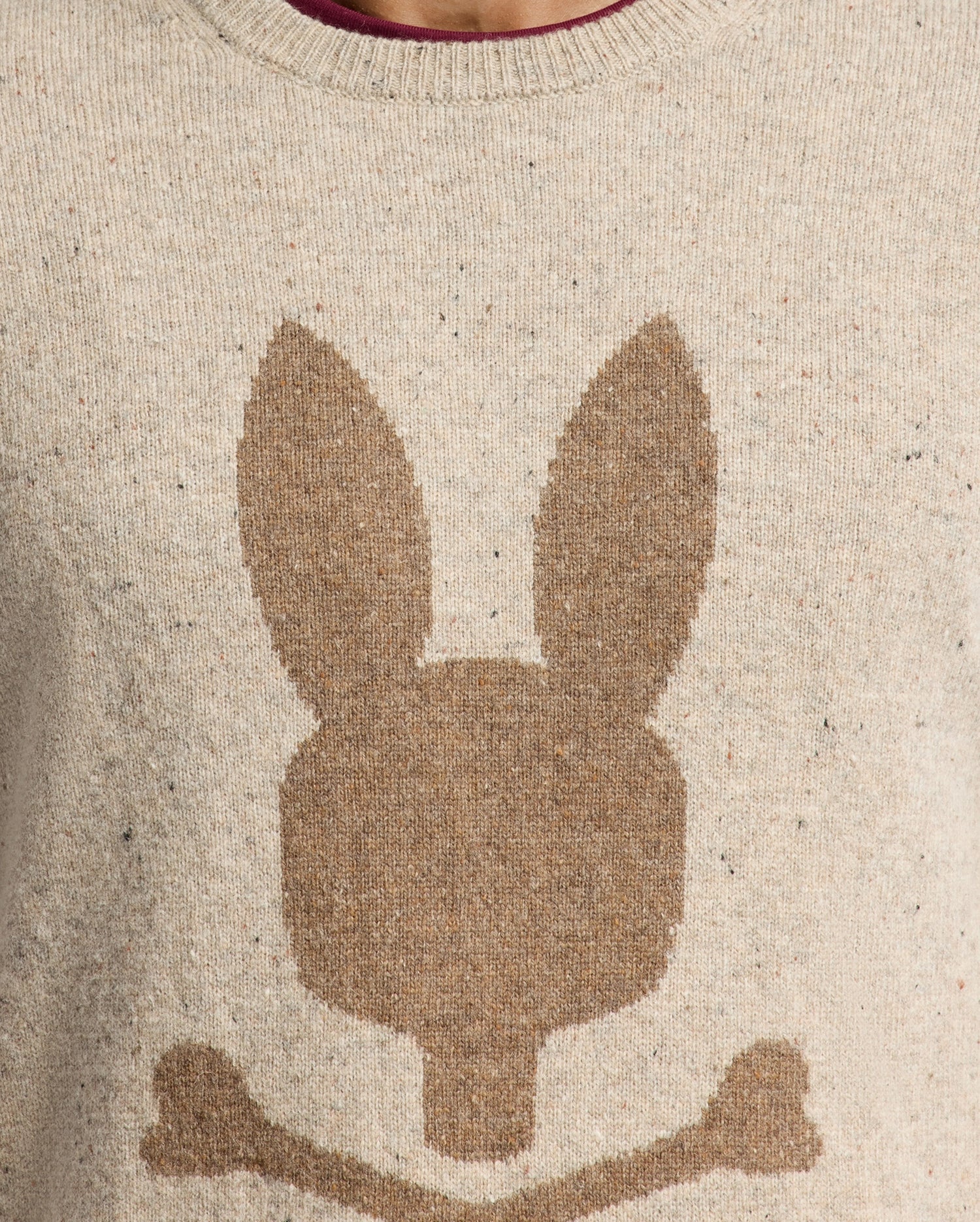 The MENS LINCOLN MERINO WOOL SWEATER - B6E831E200 by Psycho Bunny is showcased in a close-up image. This beige sweater features a brown silhouette of a stylized rabbit head with long ears above two crossed bones, highlighting the texture of the warm Merino wool knit fabric.