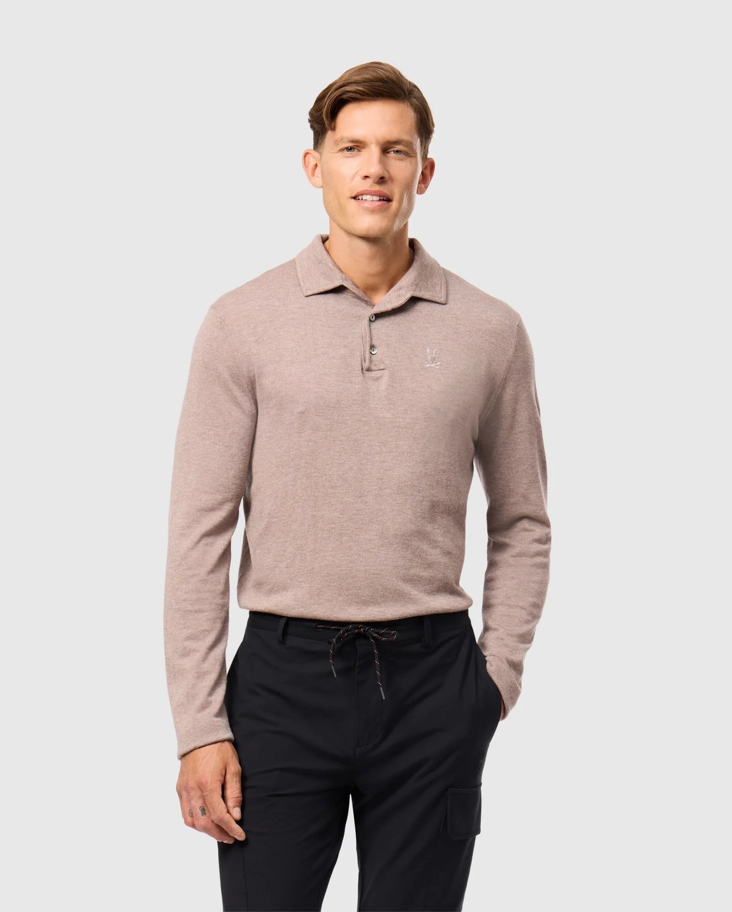 Wearing the light brown Psycho Bunny MENS BRADSHAW POLO SWEATER - B6E835D200 and black pants, a person stands against a plain gray background. They have short hair, a slight smile, and their hands at their sides.