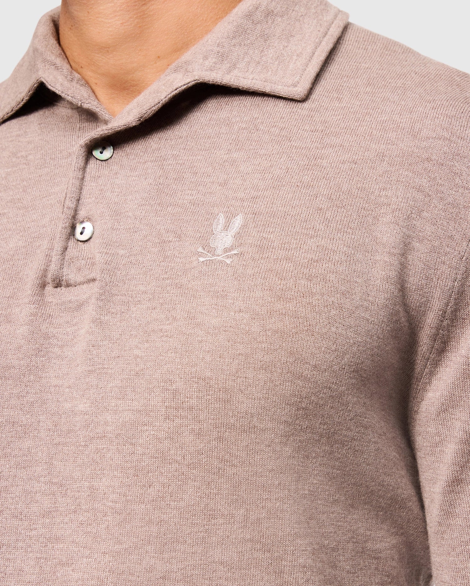 A close-up captures a person wearing the Psycho Bunny MENS BRADSHAW POLO SWEATER - B6E835D200, showcasing its distinctive rabbit logo on the chest. The luxury merino wool sweater for men features two buttons undone at the collar, emphasizing its soft texture and elegance. The focus remains on the upper torso against a plain background.