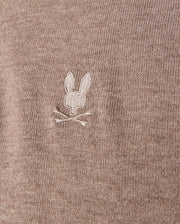 Detailed view of the brown MENS BRADSHAW POLO SWEATER by Psycho Bunny, crafted from luxurious merino wool. It features a small, white embroidered bunny head with crossed bones beneath it, adding a subtle detail to the fine texture of the fabric.