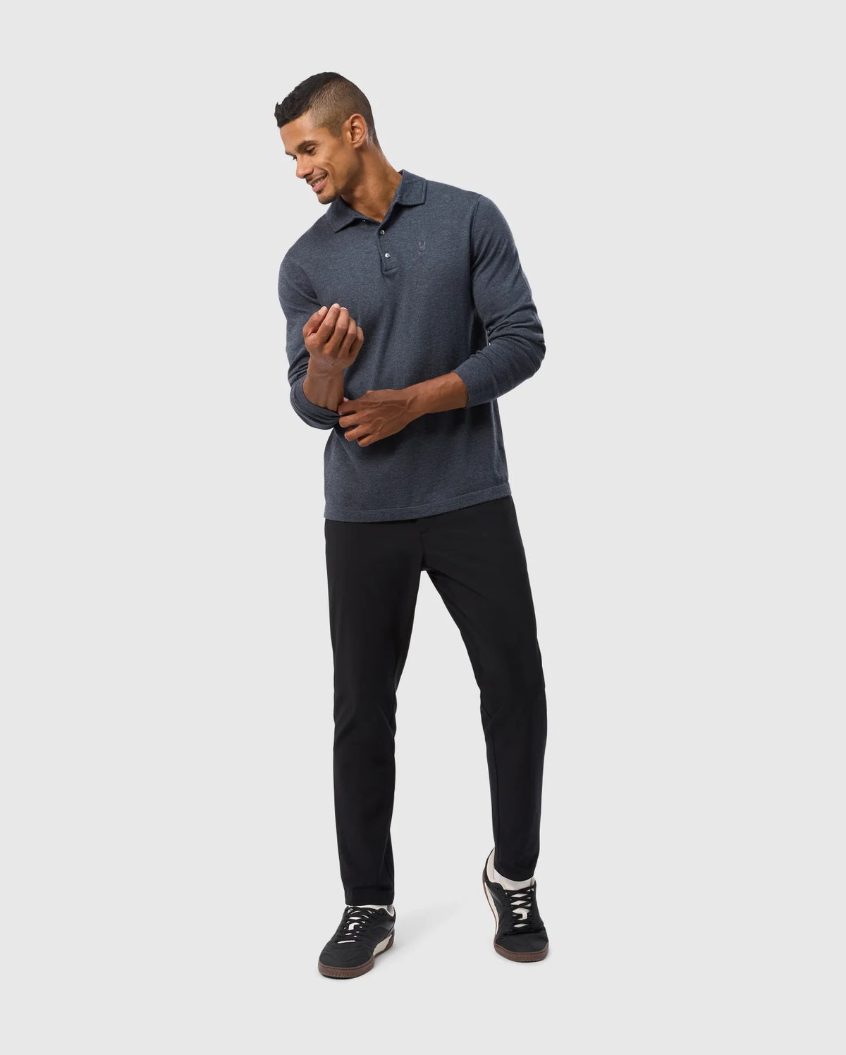 A man in a relaxed pose, wearing the Psycho Bunny MENS BRADSHAW POLO SWEATER - B6E835D200, paired with black pants, adjusts his sleeve while looking down. He has short hair and a subtle smile, standing against a simple light grey background that highlights his comfort and style.