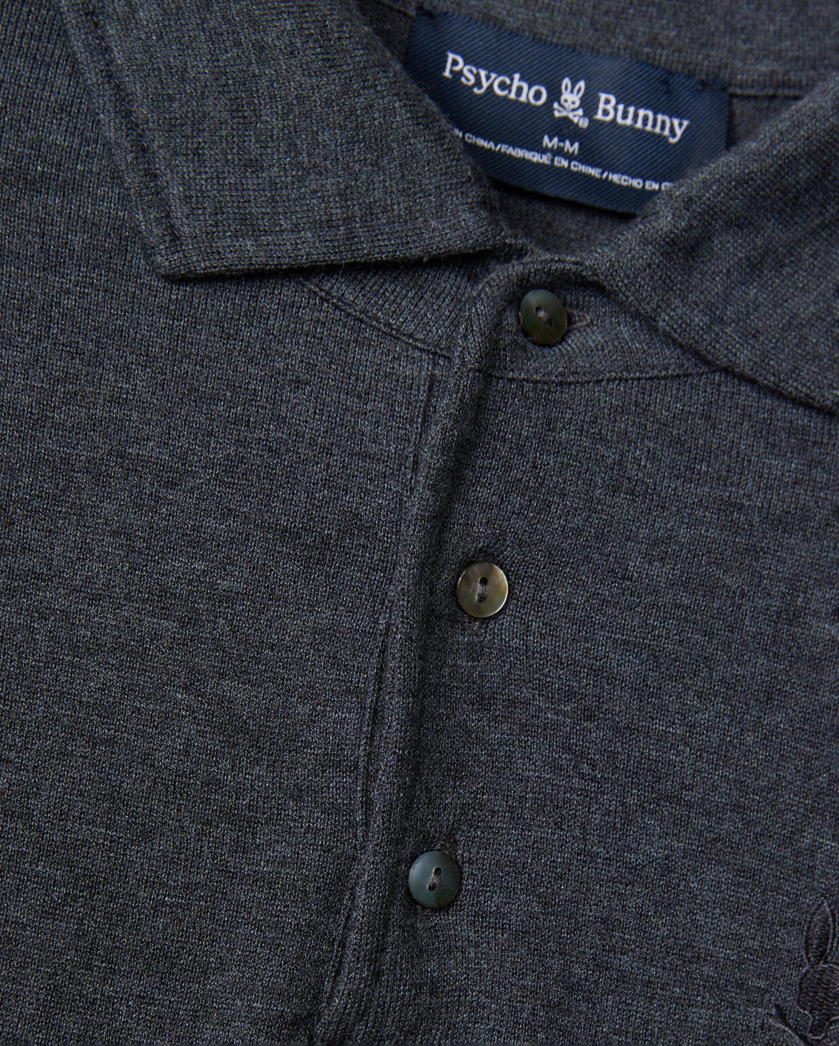 A close-up of the dark gray Men's Bradshaw Polo Sweater (B6E835D200) showcases its merino wool fabric, complete with buttons and a collar. The Psycho Bunny brand label is prominently displayed at the neck, featuring the iconic bunny logo that accentuates its luxurious appeal.
