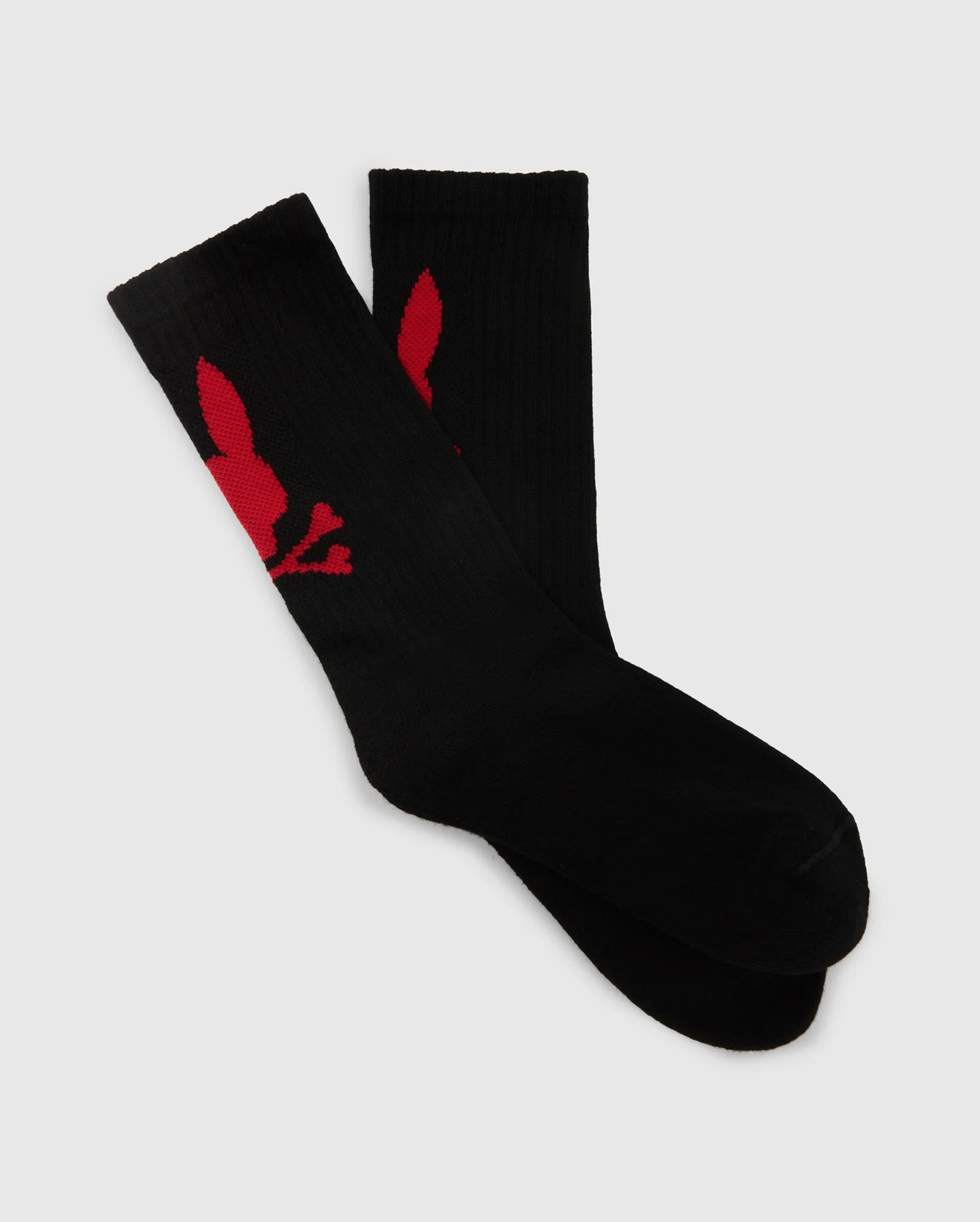 Introducing the MENS SPORT SOCK BOX SET - B6F268E200 by Psycho Bunny: This pair of men's sport socks, crafted from luxurious Peruvian Pima Cotton, ensures exceptional softness. They feature a distinct red silhouette of a bunny head with ears positioned diagonally on each black sock, set against a light gray background.