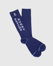 A pair of navy blue dress socks from Psycho Bunny, crafted from Pima cotton, feature "PSYCHO BUNNY" text in white and a small bunny logo with crossbones. Lighter blue stitching accents the heels and toes, making them perfect for any stylish look. Part of the MENS 3 PACK DRESS SOCK - B6F504F200 set.