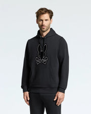 A person stands wearing the MENS ALEXANDER HOODIE - B6H154E200 from Psycho Bunny, which features a bunny and crossbones graphic on the front. The hoodie includes a front pocket, and the wearer complements it with matching black pants. The background is plain and light-colored.