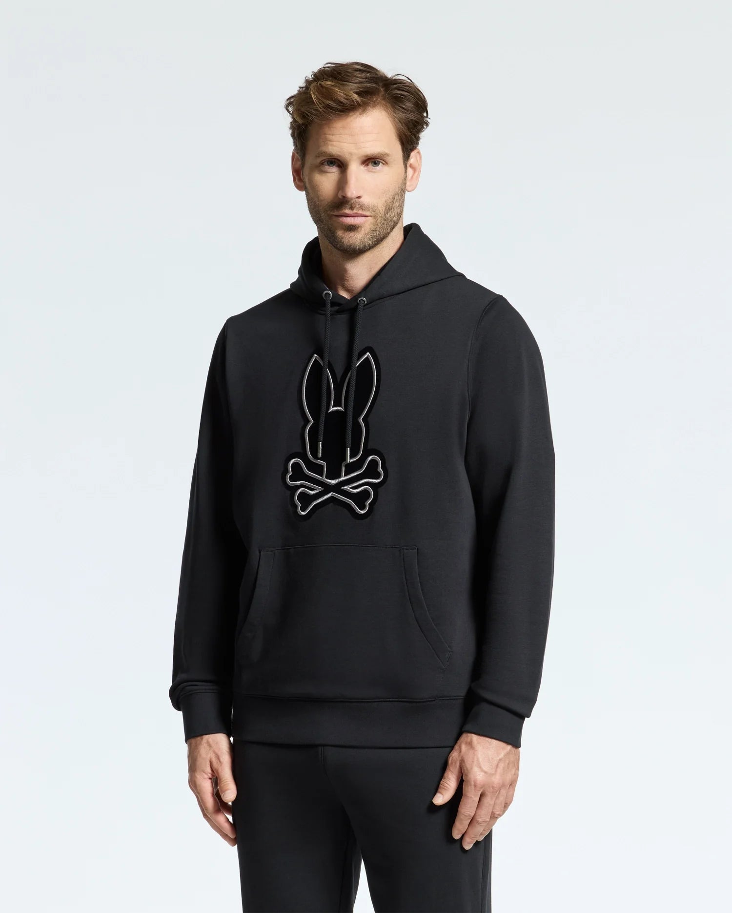 A person stands wearing the MENS ALEXANDER HOODIE - B6H154E200 from Psycho Bunny, which features a bunny and crossbones graphic on the front. The hoodie includes a front pocket, and the wearer complements it with matching black pants. The background is plain and light-colored.