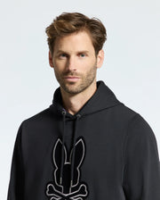 A man with brown hair and facial hair wears the comfortable Psycho Bunny men's Alexander hoodie, adorned with a bunny and skull design. The black, soft touch men's hooded sweatshirt stands out against the plain white background.