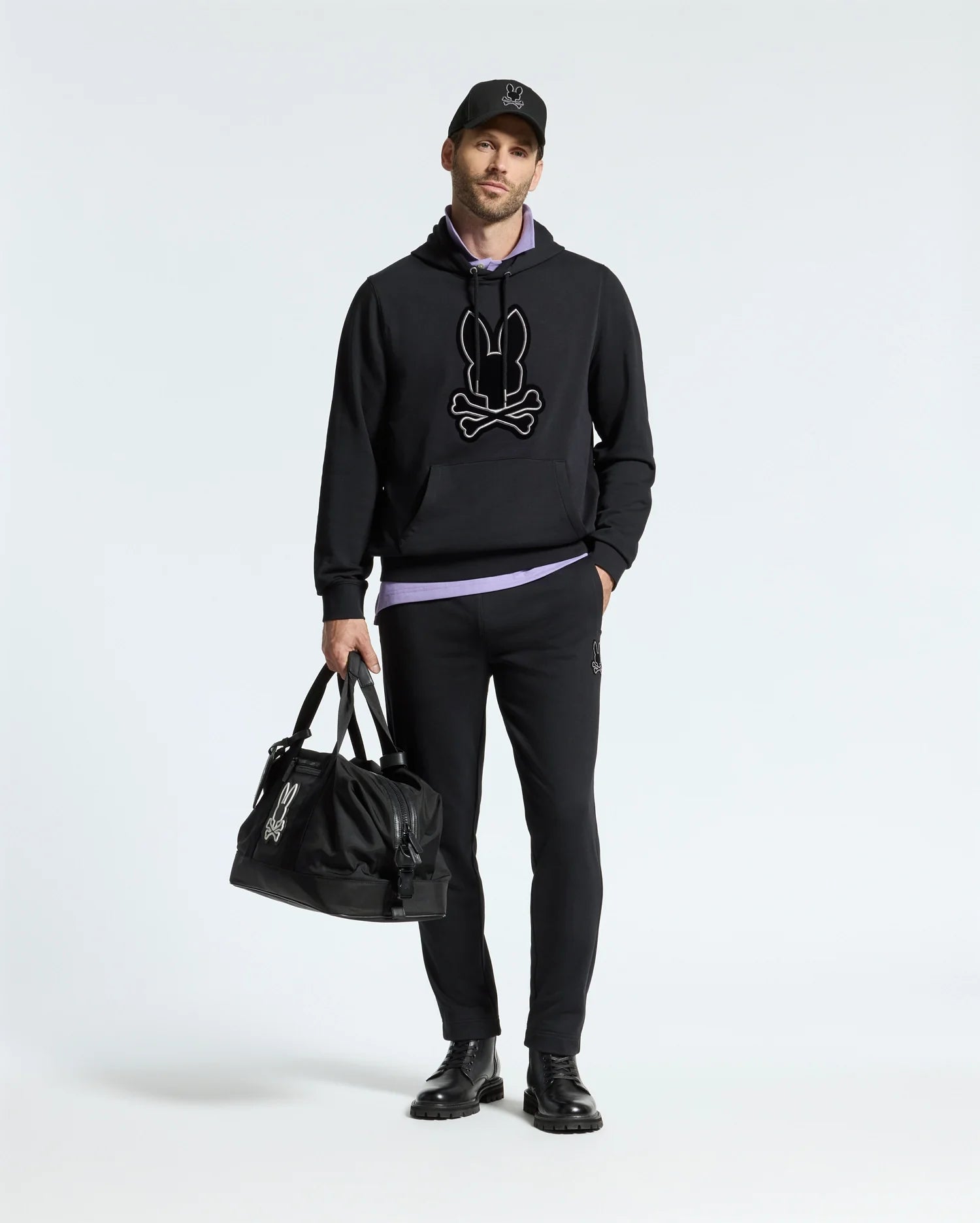 A person wearing a black Psycho Bunny MENS ALEXANDER HOODIE - B6H154E200, black pants, and black shoes stands holding a matching duffle bag against a plain white background.