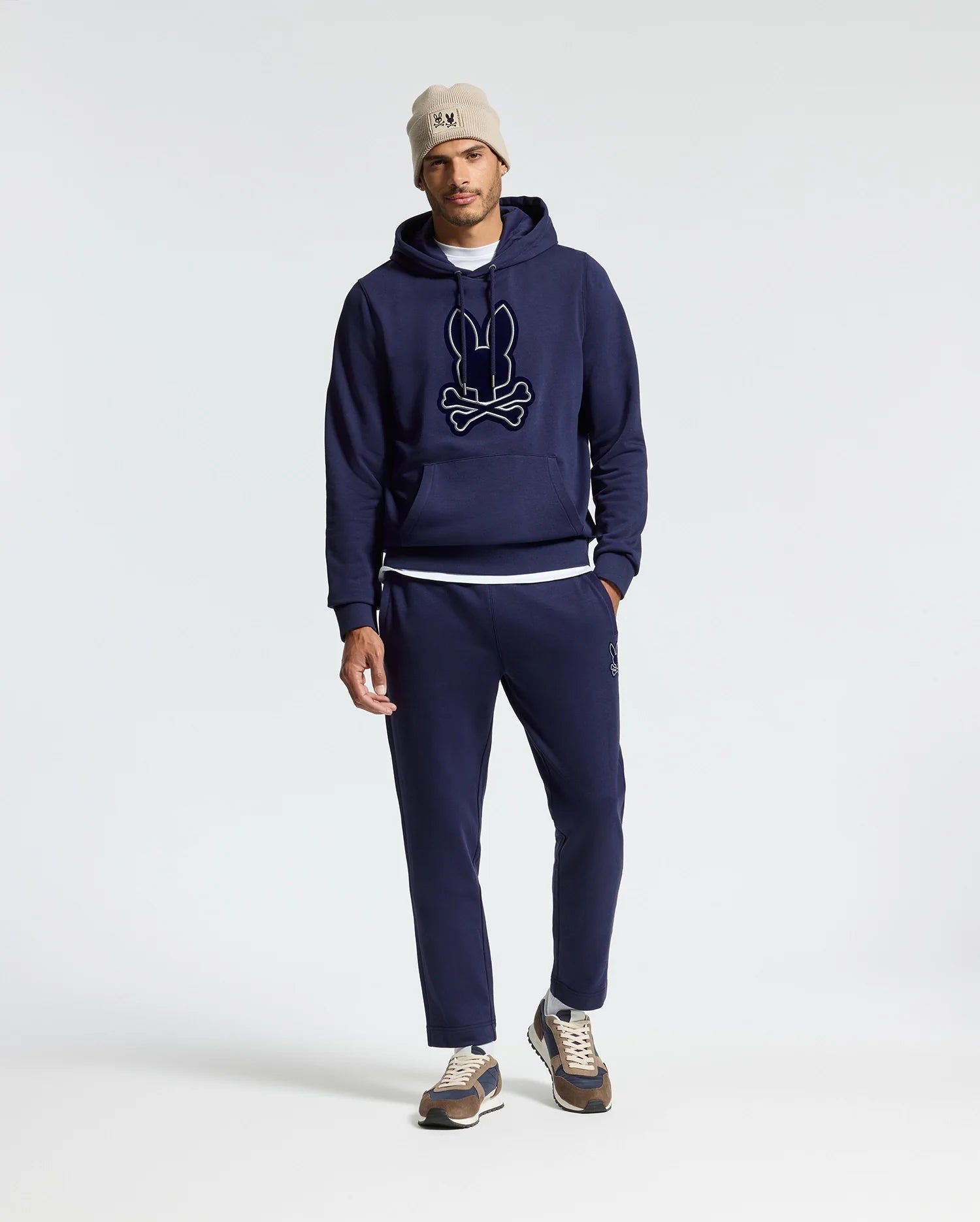 A person wearing a Psycho Bunny MENS ALEXANDER HOODIE - B6H154E200 along with matching jogger pants, with a white T-shirt slightly visible, a beige beanie, and sneakers. The comfortable men's hoodie showcases a graphic design on the front. The outfit is displayed against a simple, light-colored background.
