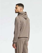A man stands sideways against a plain white background, wearing the Psycho Bunny MENS ALEXANDER HOODIE - B6H154E200 in light brown with matching sweatpants.