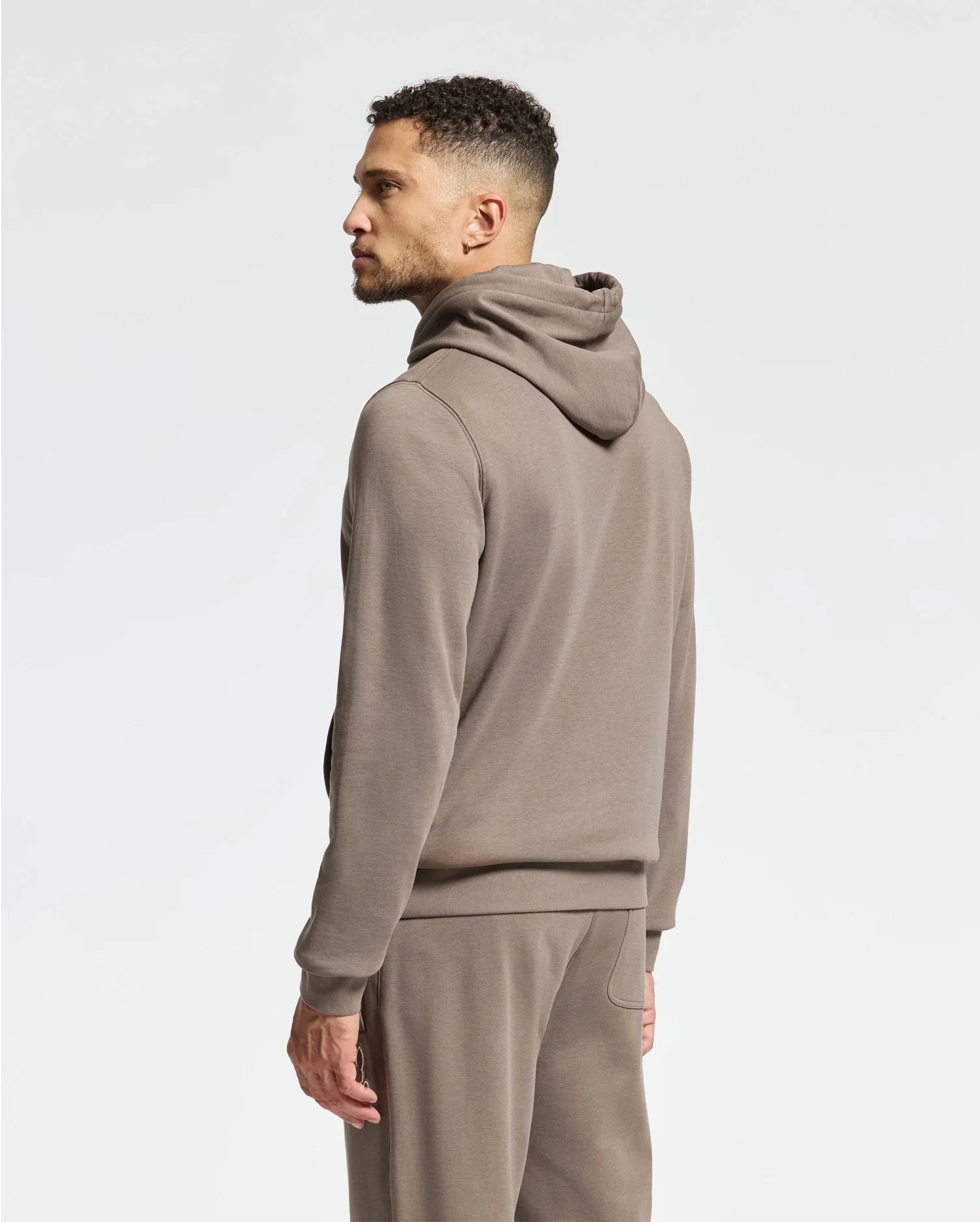A man stands sideways against a plain white background, wearing the Psycho Bunny MENS ALEXANDER HOODIE - B6H154E200 in light brown with matching sweatpants.