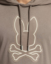 A close-up of the Psycho Bunny MENS ALEXANDER HOODIE - B6H154E200 showcases a brown pullover featuring a logo. The logo, depicting rabbit ears above crossed bones, mimics a skull and crossbones design with comfortable drawstrings visible on either side.