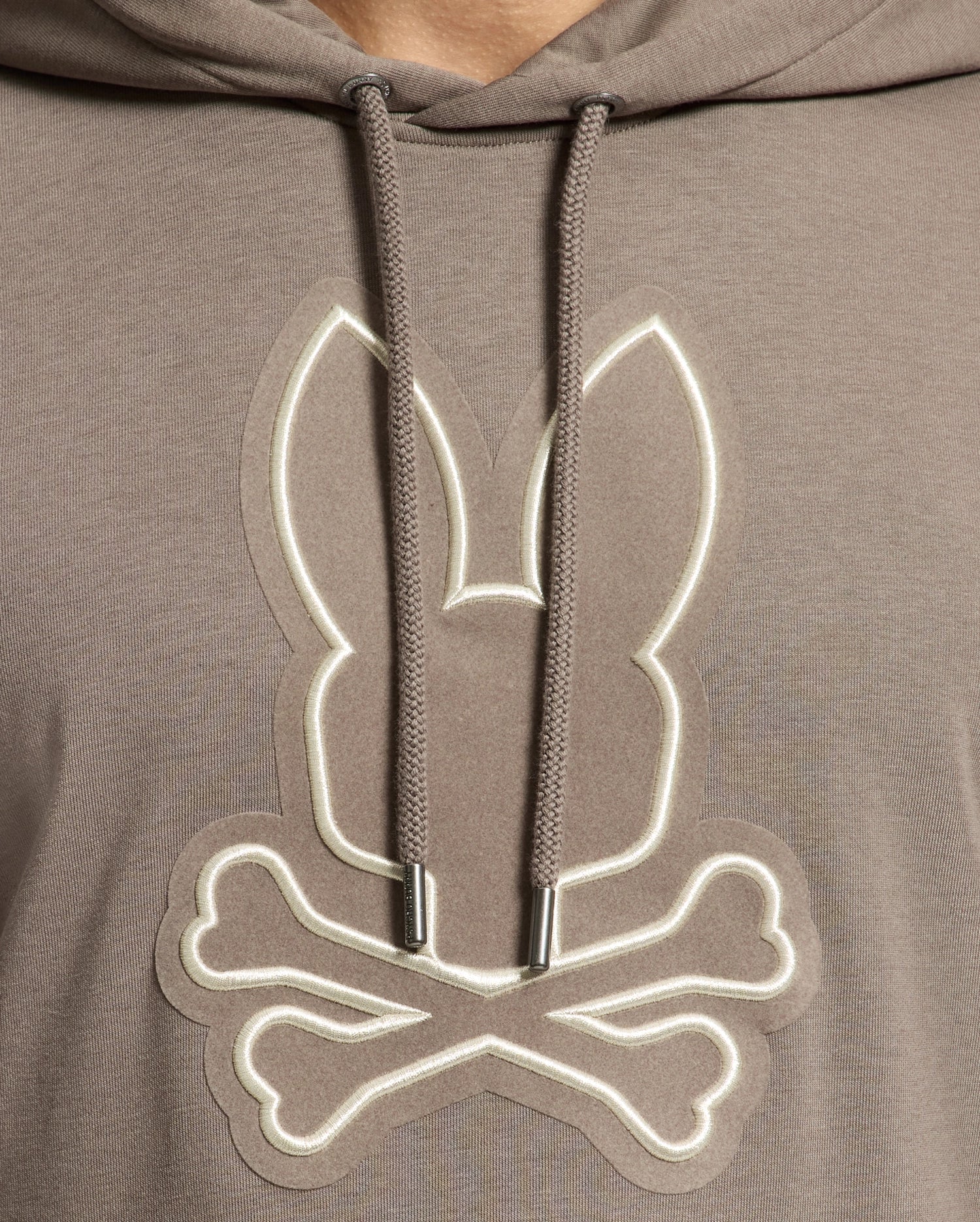A close-up of the Psycho Bunny MENS ALEXANDER HOODIE - B6H154E200 showcases a brown pullover featuring a logo. The logo, depicting rabbit ears above crossed bones, mimics a skull and crossbones design with comfortable drawstrings visible on either side.