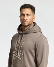 A person with short curly hair is comfortably wearing the Psycho Bunny MENS ALEXANDER HOODIE - B6H154E200, featuring a stylized rabbit and crossbones design in gray. The pullover stands out against the plain light-colored background.