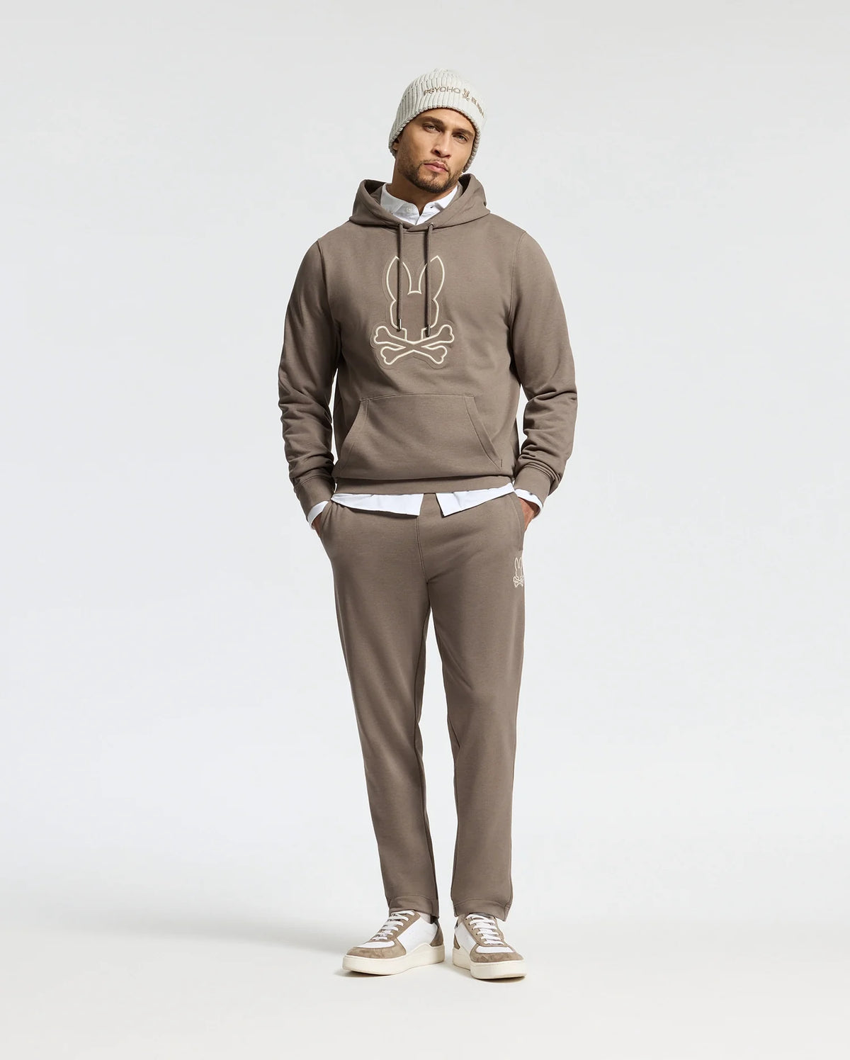 A person is dressed in a comfortable taupe Psycho Bunny MENS ALEXANDER HOODIE - B6H154E200, showcasing a cartoonish design, paired with matching sweatpants and a white shirt underneath. The outfit is completed with a light gray beanie and gray sneakers against a plain white background.