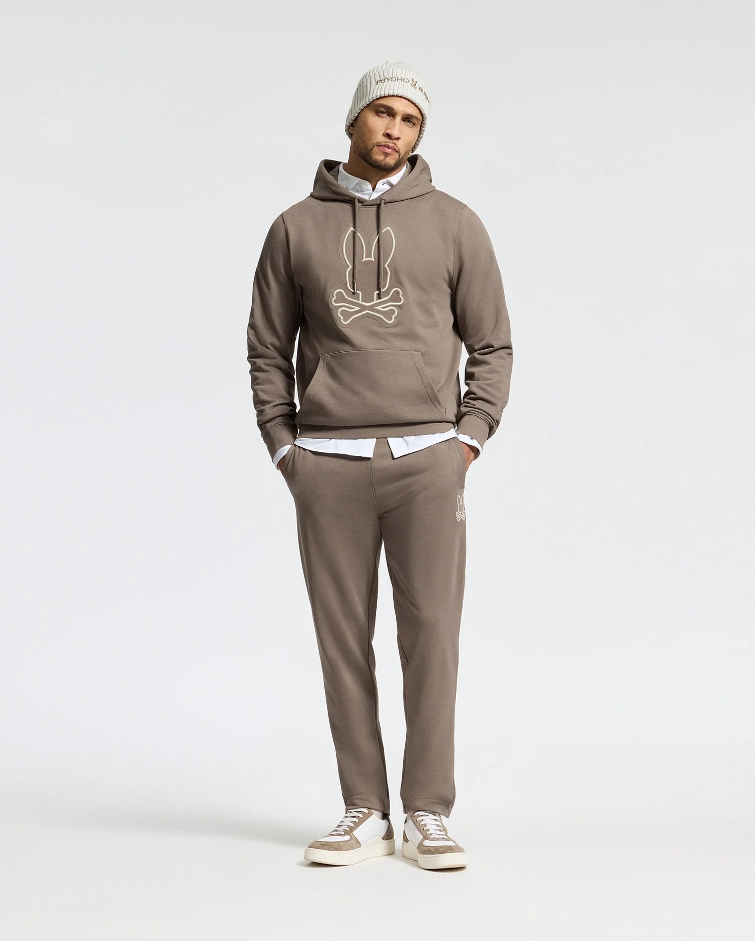 A person is dressed in a comfortable taupe Psycho Bunny MENS ALEXANDER HOODIE - B6H154E200, showcasing a cartoonish design, paired with matching sweatpants and a white shirt underneath. The outfit is completed with a light gray beanie and gray sneakers against a plain white background.