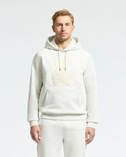 A person with short blonde hair wears the light gray Psycho Bunny MENS ELDORADO BRUSH BACK HOODIE (B6H234E200) and matching sweatpants, featuring a cartoon skull and bones design, ideal for swaddle season against a plain gray background.