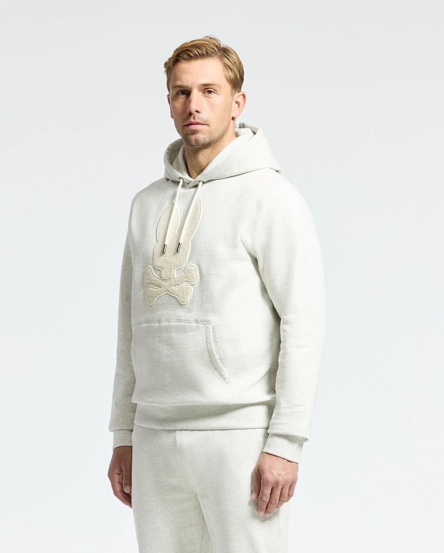 A man with light hair in a Psycho Bunny MENS ELDORADO BRUSH BACK HOODIE - B6H234E200, showcasing brush-back comfort and a cartoon design, stands against a plain background. Perfect for swaddle season, the hoodie features a cozy kangaroo pocket and pairs with his light gray pants.