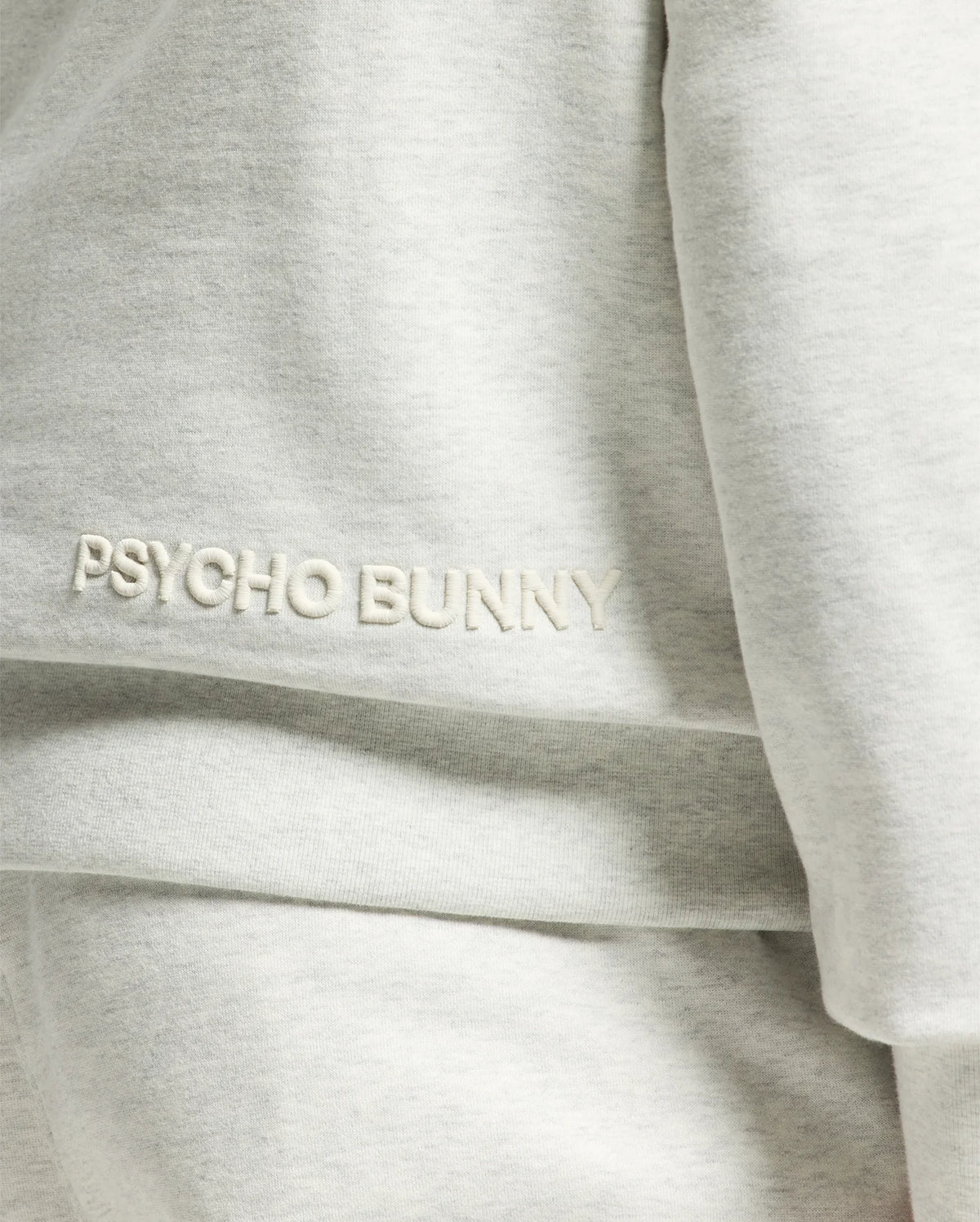 Close-up of the Psycho Bunny Mens Eldorado Brush Back Hoodie, featuring "PSYCHO BUNNY" embroidered in white on light gray, ideal for swaddle season with its cozy brush-back fabric.