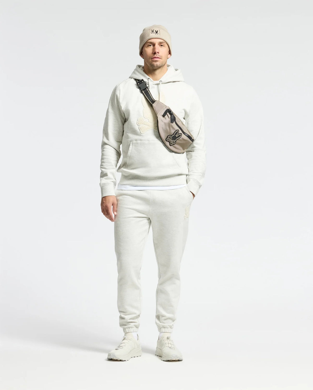 A man wears the Psycho Bunny MENS ELDORADO BRUSH BACK HOODIE in white with matching sweatpants, a beige beanie, and crossbody bag. He stands against a plain background with hands by his sides, embodying cozy brush-back comfort for swaddle season.