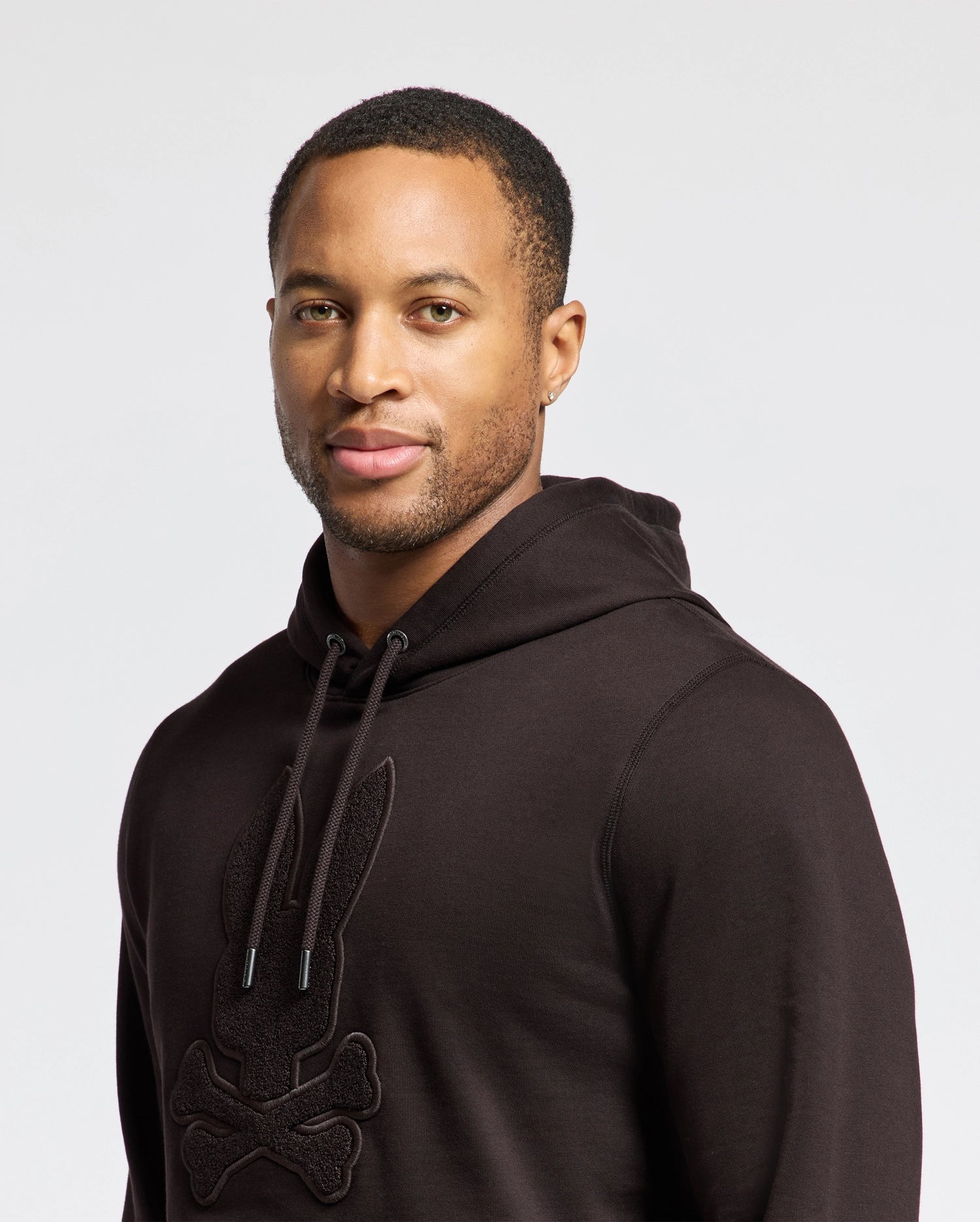 A person wears the Psycho Bunny MENS ELDORADO BRUSH BACK HOODIE (B6H234E200) featuring a stylized rabbit and crossbones design on soft brush-back cotton with a kangaroo pocket, standing against a white background.