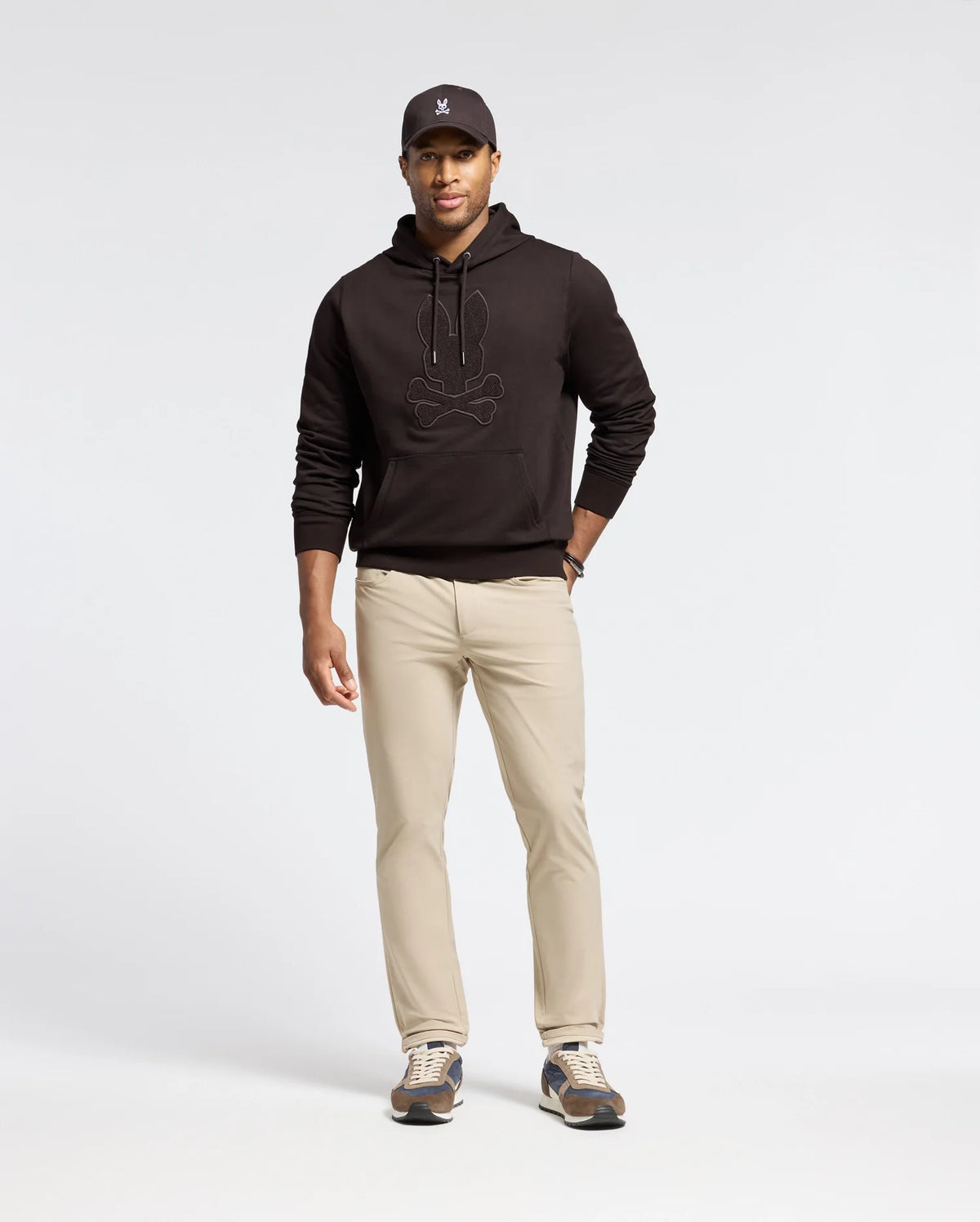A man in a Psycho Bunny MENS ELDORADO BRUSH BACK HOODIE - B6H234E200 in black, graphic beige pants, a black cap, and sneakers stands against a plain white background. The hoodie features a cozy kangaroo pocket for style and functionality.
