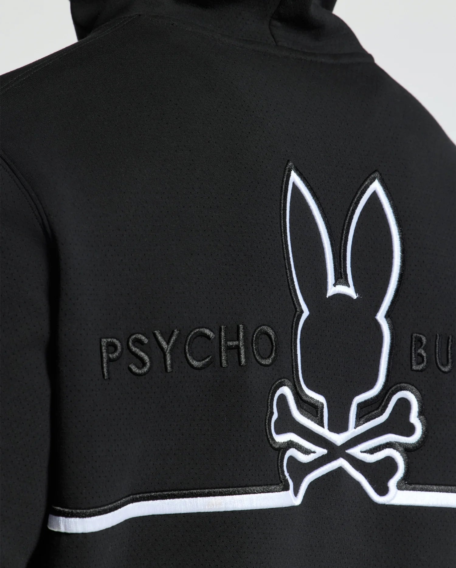 Close-up of the back of a Psycho Bunny MENS DACANO PERFORATED HOODIE in black, showcasing an embroidered bunny skull and crossbones logo with "PSYCHO BUN" text in white. It features breathable, perforated texture and a horizontal white trim below the logo.