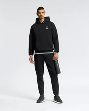 Wearing a Psycho Bunny MENS DACANO PERFORATED HOODIE in black with an embroidered logo, black joggers, and sleek black sneakers, the person stands holding a black water bottle against a plain white background.