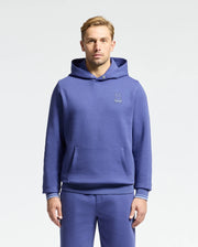 A person with short hair stands against a plain backdrop, wearing Psycho Bunny's DACANO Perforated Hoodie and matching sweatpants in blue stretch cotton-blend jersey. The hoodie displays a small embroidered rabbit logo on the left side, while their hands rest by their sides.