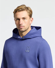 A person with short blond hair wears Psycho Bunny's Mens Dacano Perforated Hoodie (B6H239E200), a stretch cotton-blend jersey in blue featuring an embroidered bunny and crossed bones logo on the chest, against a plain white background.