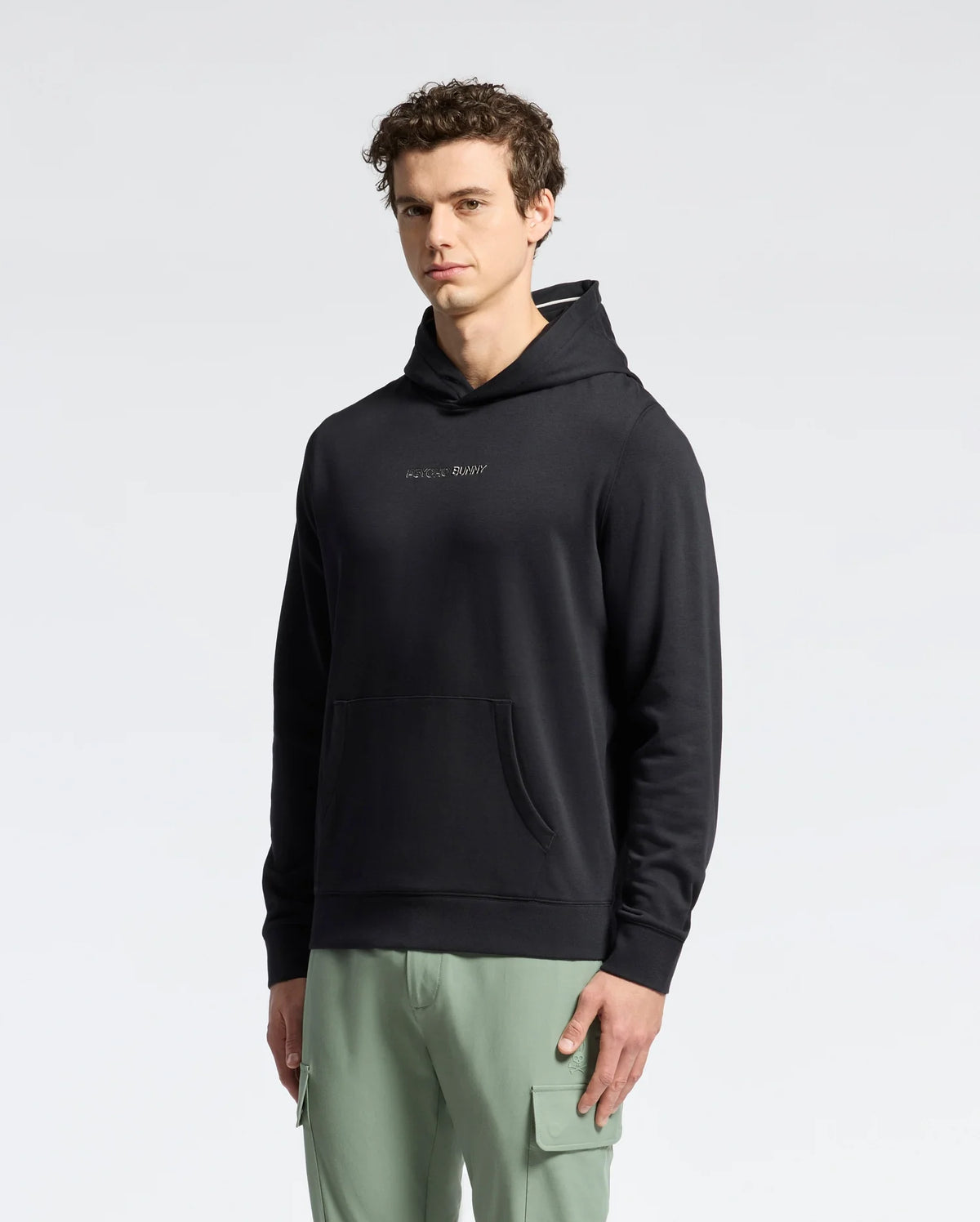 A man faces the camera in a Psycho Bunny MENS NORA CLASSIC FRENCH TERRY HOODIE - B6H434F200, featuring an embroidered chest logo, paired with light green cargo pants against a plain, light backdrop.