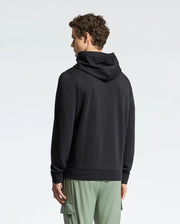 A person with curly hair is facing away, wearing a black Psycho Bunny MENS NORA CLASSIC FRENCH TERRY HOODIE and green cargo pants against a plain white background.