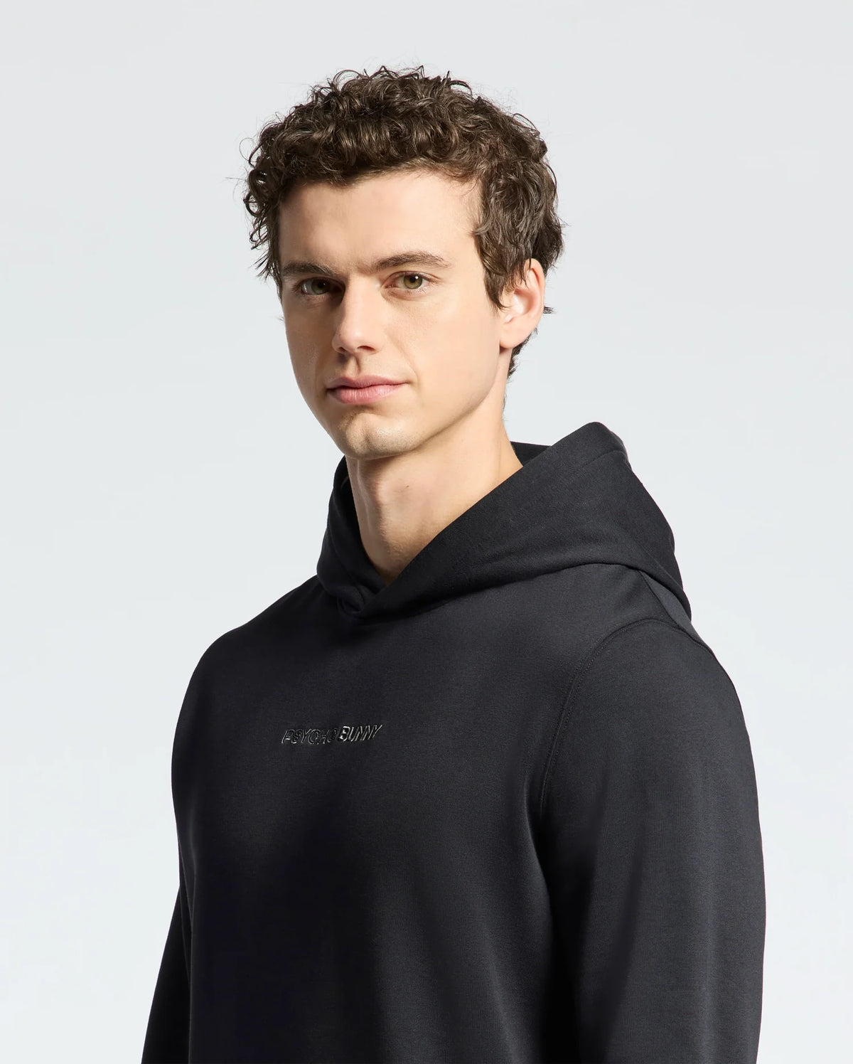 A person with curly dark hair wearing a black Mens Nora Classic French Terry Hoodie by Psycho Bunny stands against a plain light background, looking slightly to the side. The hoodie features a subtle embroidered logo across the chest.