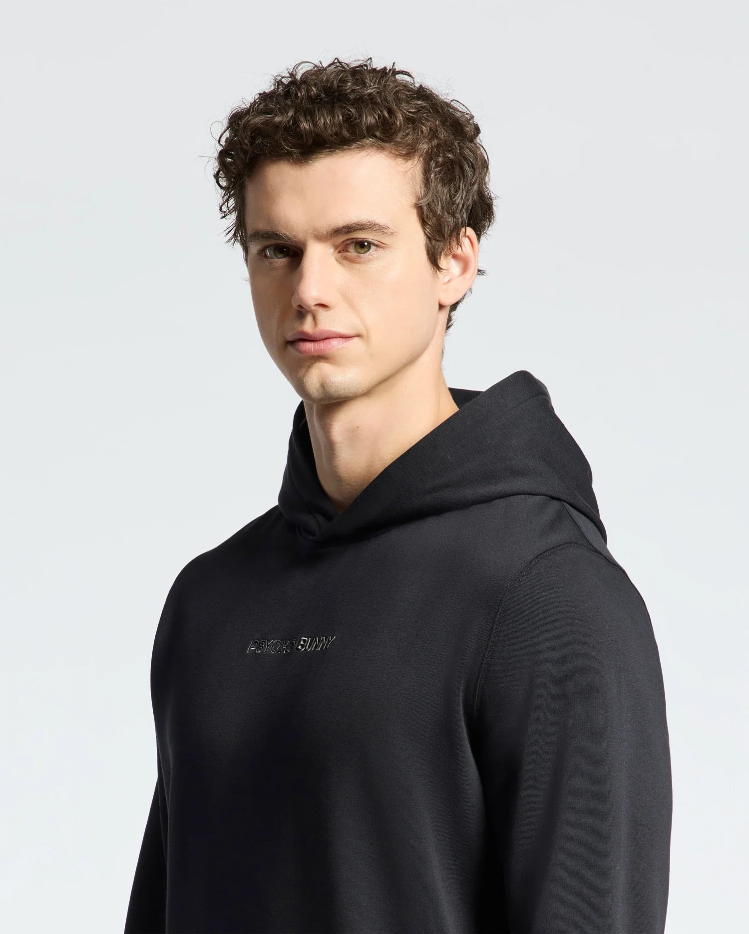 A person with curly dark hair wearing a black Mens Nora Classic French Terry Hoodie by Psycho Bunny stands against a plain light background, looking slightly to the side. The hoodie features a subtle embroidered logo across the chest.