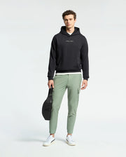 A person with curly brown hair stands against a plain background wearing light green pants, white sneakers, and a black Psycho Bunny Nora Classic French Terry Hoodie. They hold a black bag in one hand, completing their casual look.