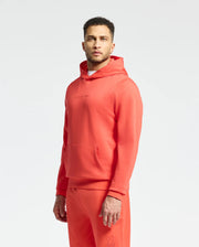 Someone is wearing a Psycho Bunny Nora Hoodie and sweatpants in coral, made from cozy French terry. The hoodie has an embroidered wordmark on the chest, while the pants display a subtle logo near the pocket. A plain white background enhances this stylish ensemble.