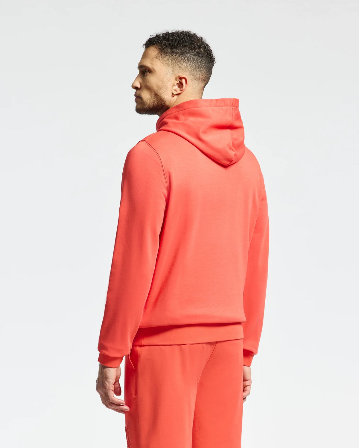 A man with short curly hair, facing left, wears a vibrant Psycho Bunny MENS NORA CLASSIC FRENCH TERRY HOODIE (B6H434F200) and matching joggers. The bright red set features an embroidered wordmark on the chest, against a plain white background.