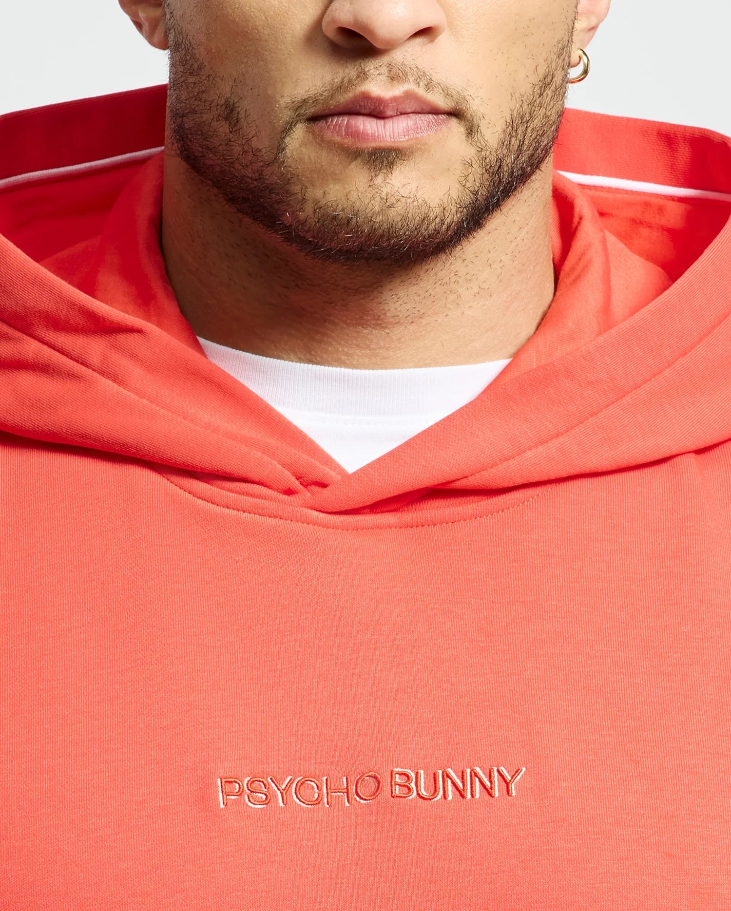 A person wearing the Men's Nora Classic French Terry Hoodie in coral from Psycho Bunny, featuring an embroidered wordmark. The hoodie, crafted from soft French terry fabric, includes a white hood lining. The background is plain white.