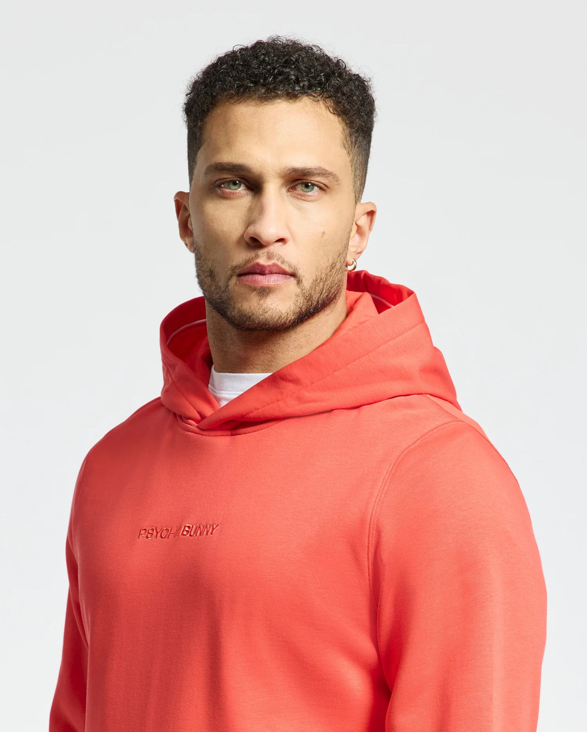 A man with short curly hair and a beard wears the coral Psycho Bunny MENS NORA CLASSIC FRENCH TERRY HOODIE - B6H434F200, showcasing an embroidered wordmark, as he stands against a plain white background.