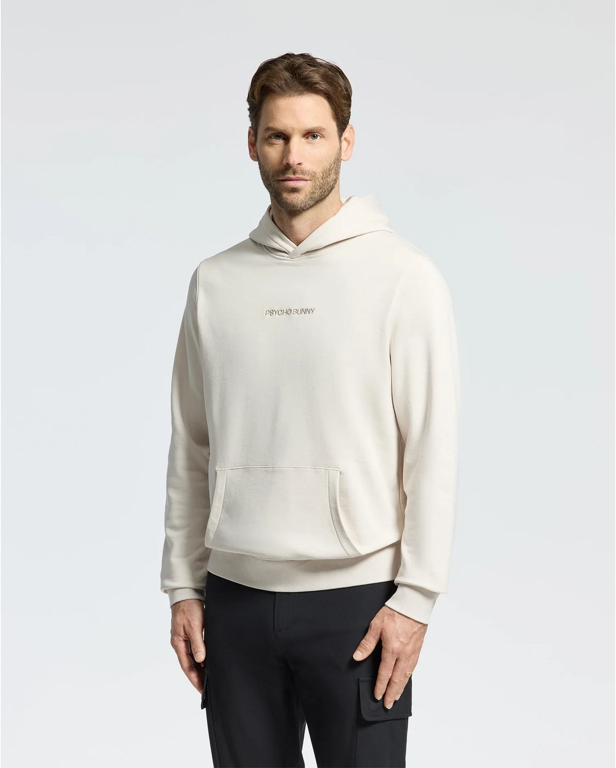 A man with medium-length brown hair and a beard wears the light beige Psycho Bunny Mens Nora Classic French Terry Hoodie (B6H434F200) featuring an "Apres Sunday" embroidered logo on the chest. He stands against a plain white background, hands relaxed by his sides.