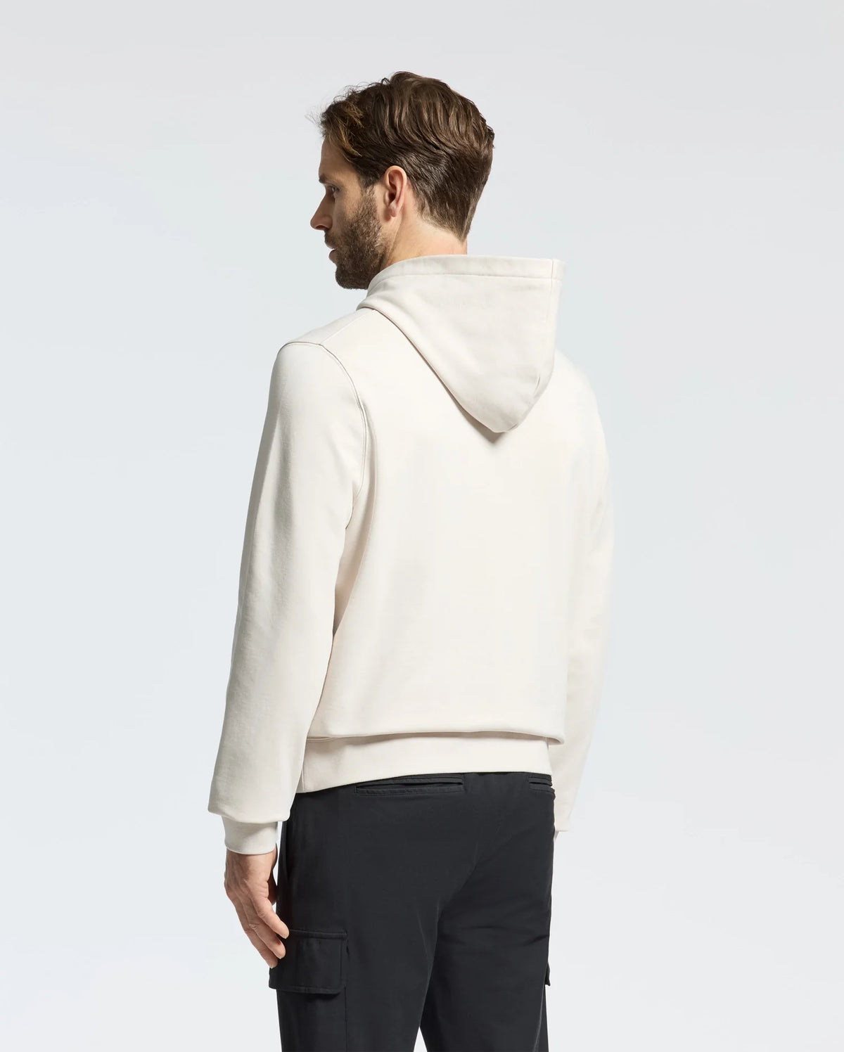 A man with short hair, seen from the side and slightly behind, wears a Psycho Bunny MENS NORA CLASSIC FRENCH TERRY HOODIE—B6H434F200. The light-colored design features a subtle embroidered logo. He pairs it with black pants against a plain white background, while the hood lays flat on his back.