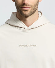 A bearded man is wearing a cream MENS NORA CLASSIC FRENCH TERRY HOODIE by Psycho Bunny with "PSYCHO BUNNY" embroidered on the front against a plain white background.
