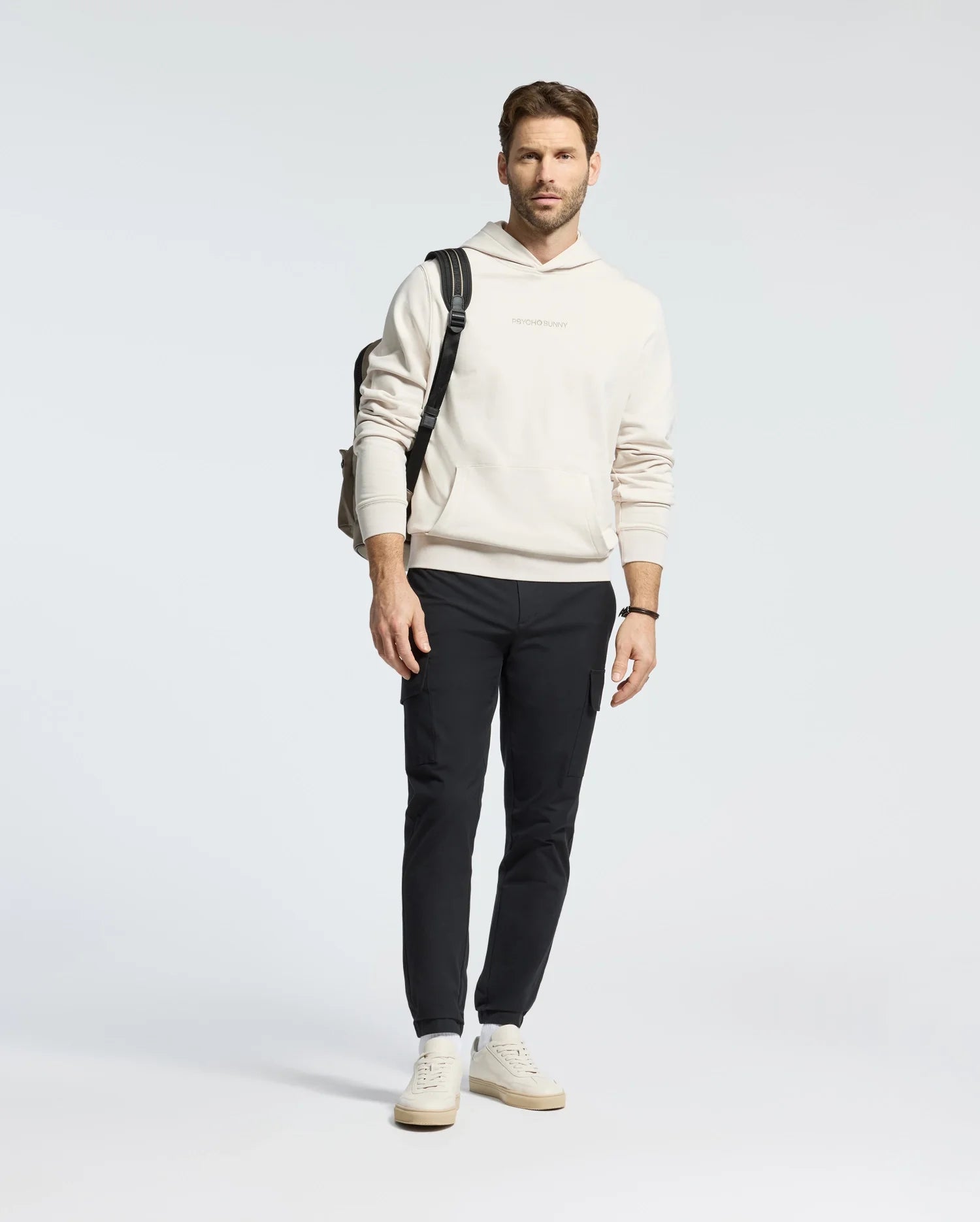 A man in a light Psycho Bunny Mens Nora Classic French Terry Hoodie (B6H434F200), black pants, and white sneakers stands against a plain background. A backpack is slung over one shoulder as he looks forward with a neutral expression.