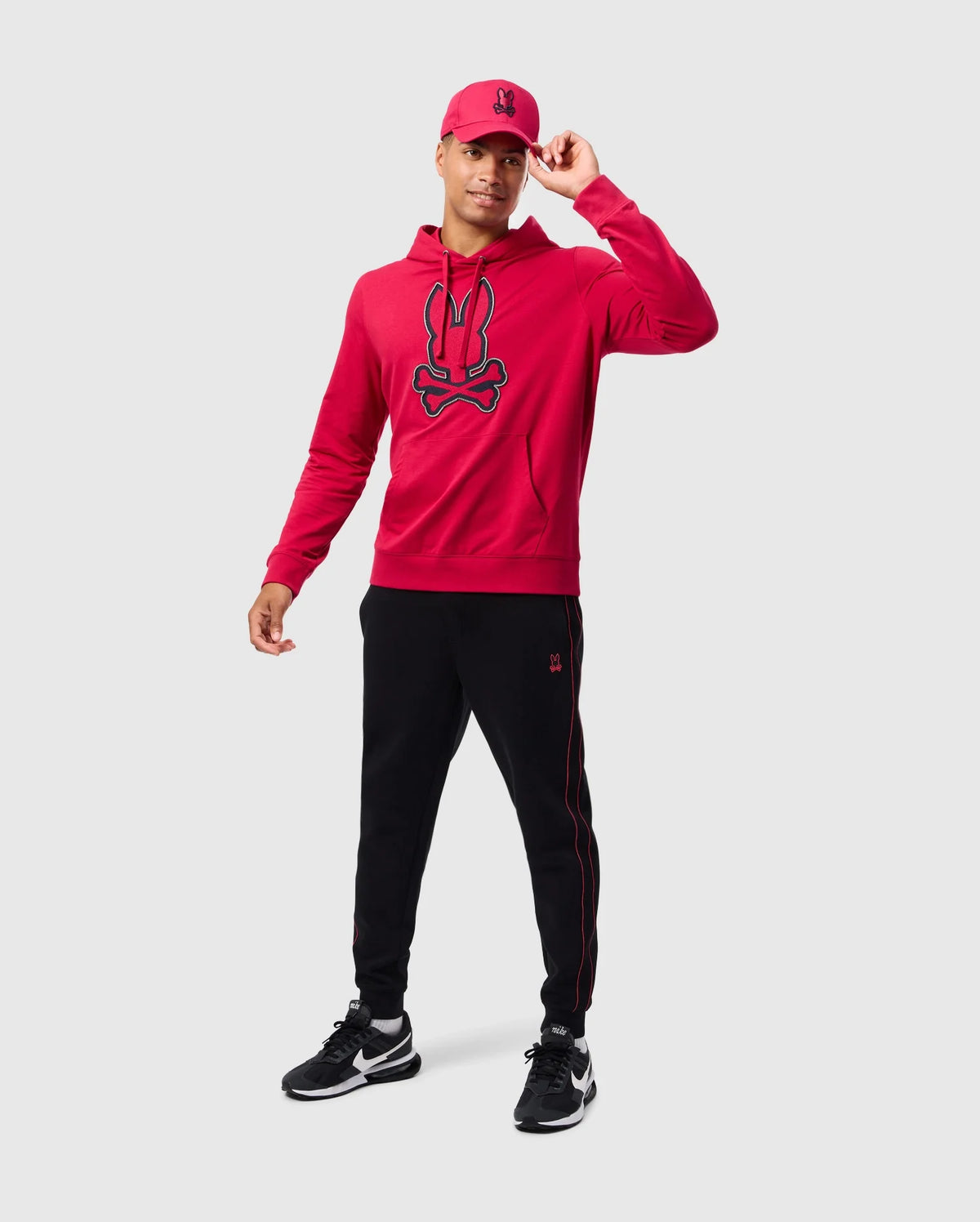 A person is donning the MENS WALTER LIGHTWEIGHT HOODIE - B6H476D200 by Psycho Bunny, a vibrant red French terry piece with a stylized logo. They pair it with black pants featuring red stripes and complete their look with black and white sneakers and a matching red cap, all set against a plain gray backdrop.