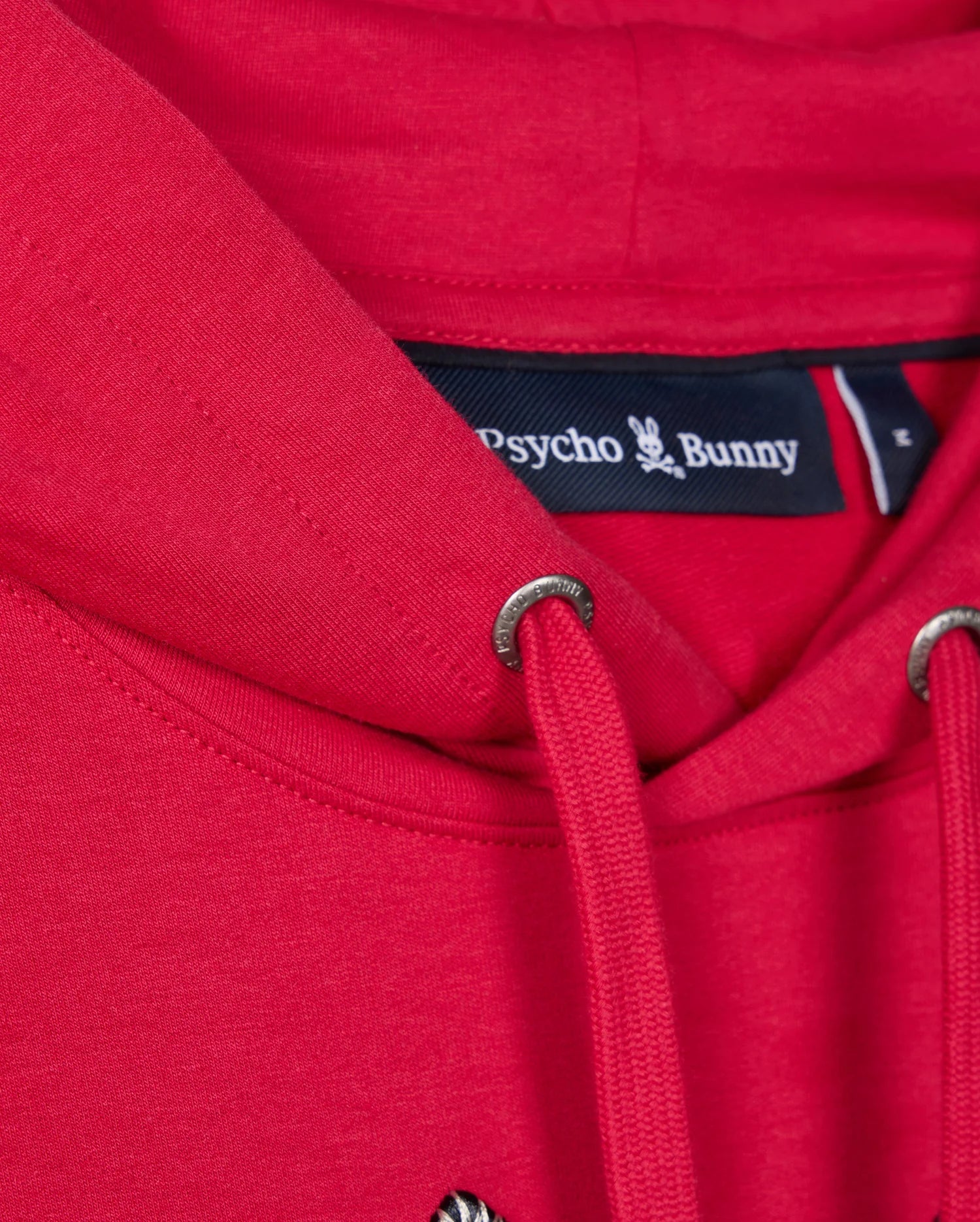A close-up of the red MENS WALTER LIGHTWEIGHT HOODIE - B6H476D200 by Psycho Bunny showcases the distinctive label at the neckline. The drawstrings with metallic eyelets contrast beautifully against the soft and comfortable Micro French Terry fabric.