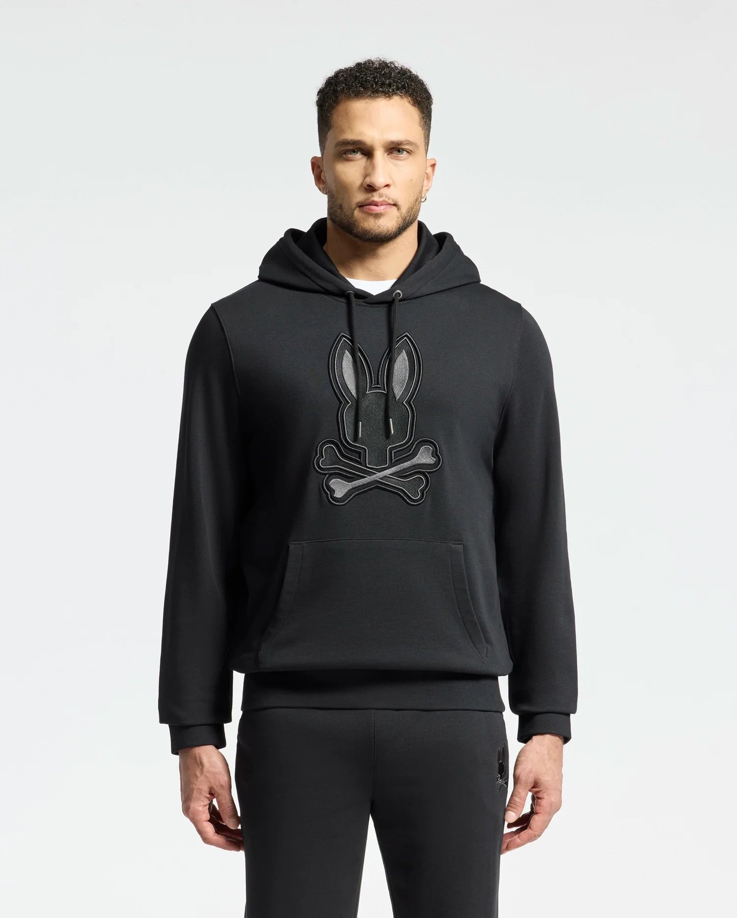 A man in an elegant Psycho Bunny MENS NEW CASTLE FRENCH TERRY HOODIE - B6H517F200, showcasing a stylized rabbit and bones design on the front, stands against a plain light background.
