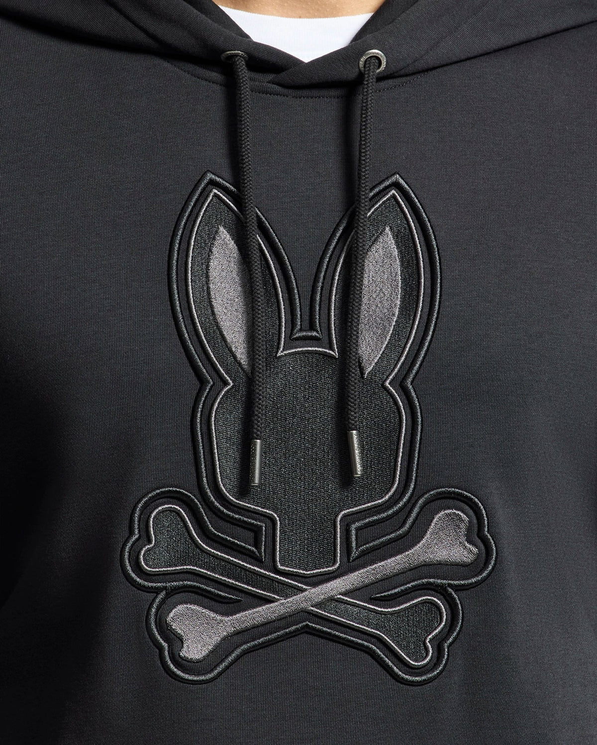 A close-up of Psycho Bunny's MENS NEW CASTLE FRENCH TERRY HOODIE (B6H517F200): an elegant black hoodie with a soft French terry fabric, showcasing a stylized embroidered bunny head and crossbones motif in gray shades on the front.