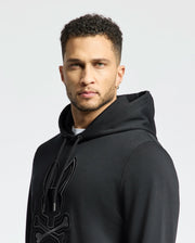 A man wearing a Psycho Bunny Men's New Castle French Terry Hoodie (B6H517F200) in black with a rabbit skull and crossbones design, gazes directly at the camera. He has short curly hair and an earring, set against a plain white background.